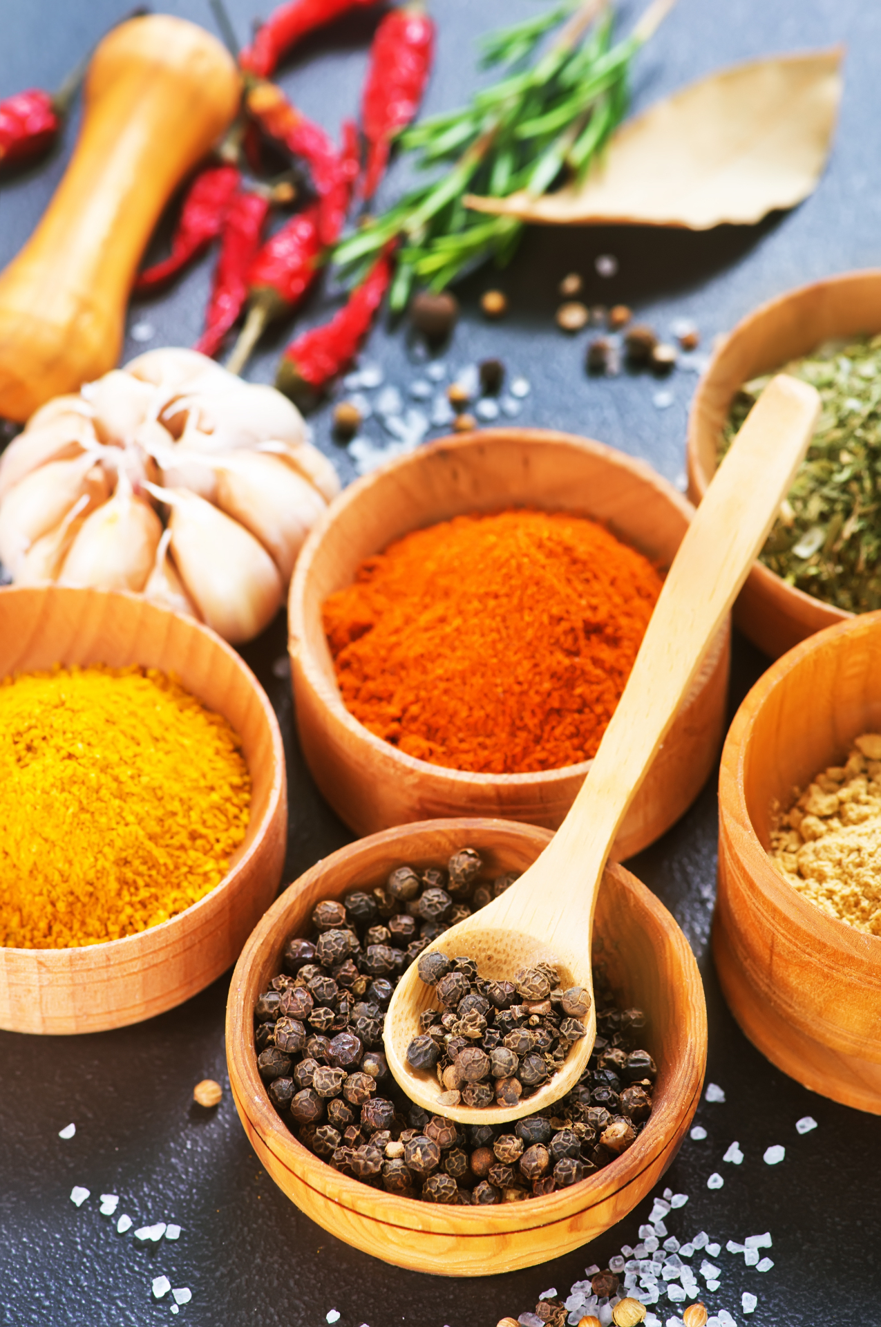 Bowls of vibrant spices, fresh herbs and peppercorns on a dark surface
